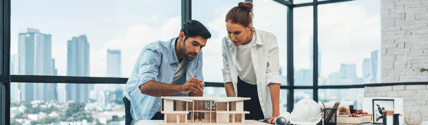 Duas pessoas analisando uma maquete arquitetônica sobre uma mesa, em um escritório com grandes janelas. Na mesa há capacete de segurança, rolos de papel e materiais de desenho.