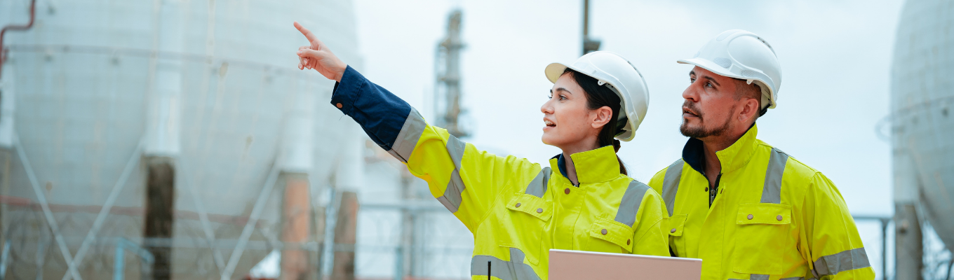Dois profissionais com capacetes e coletes de segurança analisando uma área industrial com grandes tanques esféricos ao fundo, um deles segurando um laptop e apontando para a estrutura.
