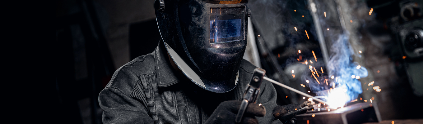Pessoa utilizando máscara de solda enquanto realiza soldagem, com faíscas e luz intensa saindo do ponto de trabalho.