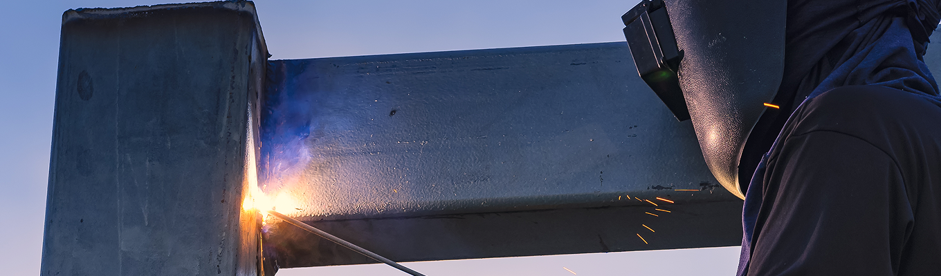 Pessoa utilizando equipamento de proteção para soldagem enquanto realiza solda utilizando eletrodo, produzindo faíscas e brilho intenso no ponto de contato