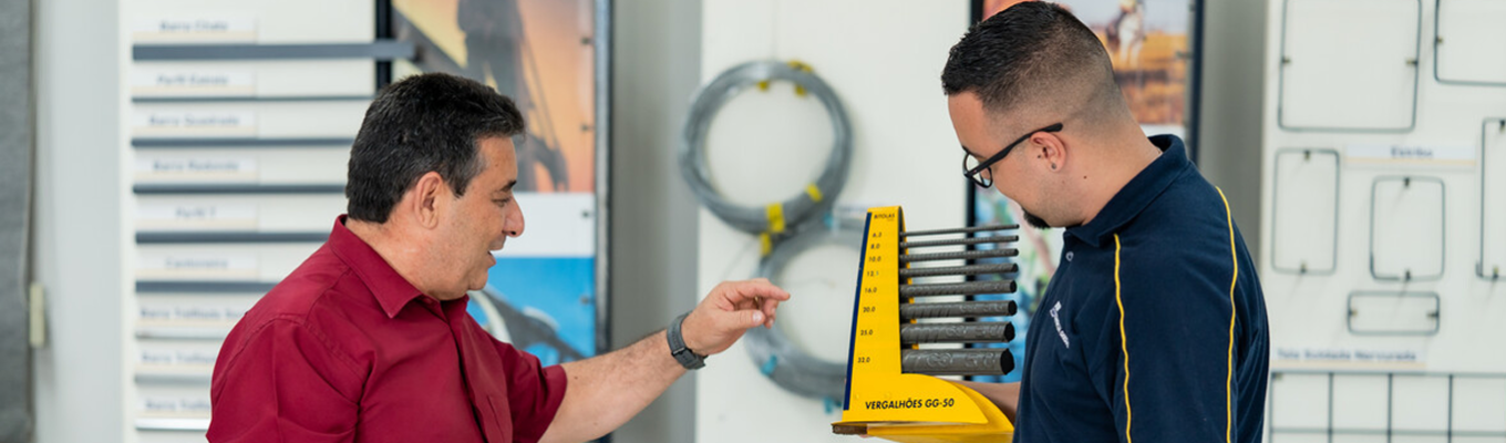 Duas pessoas interagem enquanto uma delas segura um mostruário amarelo com vergalhões de aço Gerdau em um ambiente de demonstração de produtos.