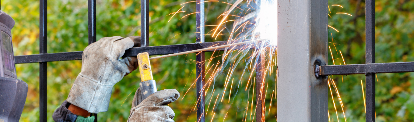 Pessoa usando luvas de proteção solda as barras de um portão metálico à sua estrutura, com faíscas visíveis, em ambiente externo com árvores ao fundo.