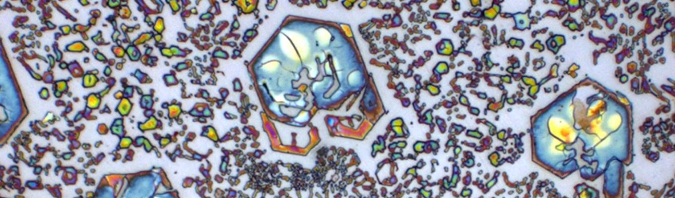 Imagem microscópica de metal com formas geométricas coloridas e partículas espalhadas, formando padrões visuais abstratos.