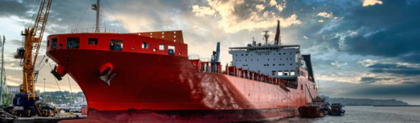 Navio de carga vermelho em estaleiro com guindastes e estrutura industrial ao fundo