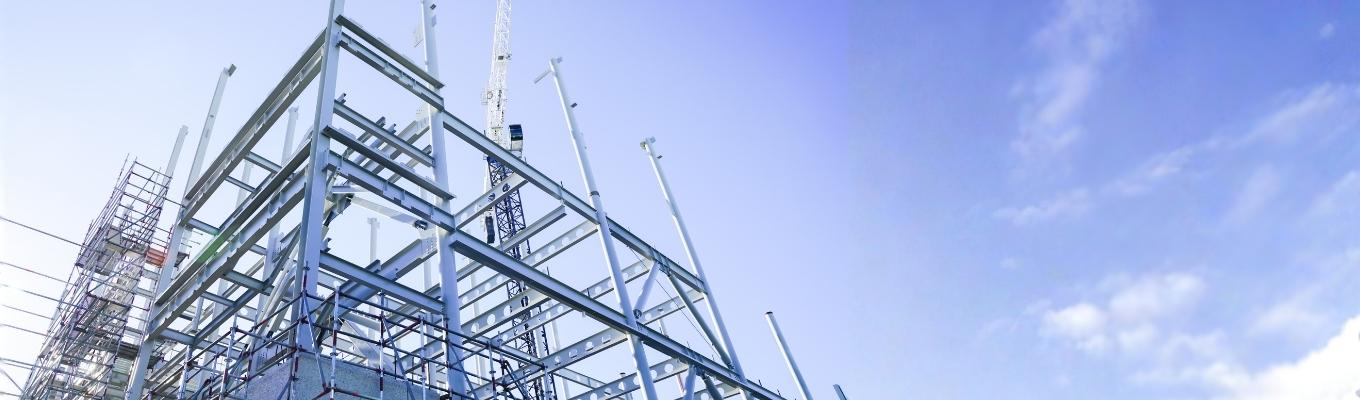 Estrutura metálica em construção com pilares e vigas de aço, andaimes e grua ao fundo.