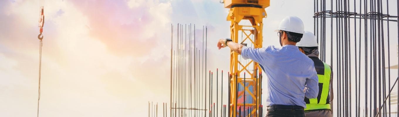 Dois profissionais usando capacetes de segurança estão em um canteiro de obras, observando e apontando para a estrutura de uma construção. No local há vergalhões verticais expostos e uma grande grua amarela ao fundo. A cena ocorre ao ar livre, com iluminação natural suave, sugerindo início ou fim de dia.