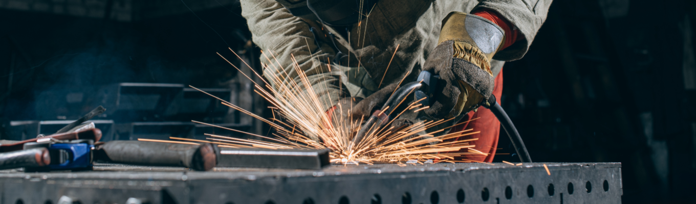 Profissional de serralheria utilizando ferramenta elétrica para soldar uma peça de metal, com muitas faíscas sobre uma superfície de aço em ambiente industrial.
