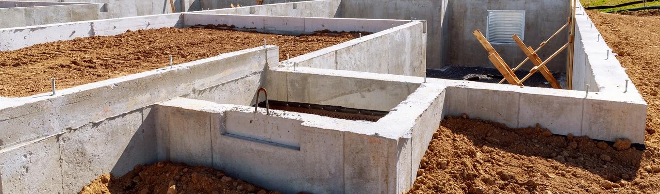 Estrutura de fundação de uma casa em construção, composta por paredes de concreto formando diversos compartimentos subterrâneos, com terra ao redor e dentro das áreas delimitadas. No ambiente externo, há um terreno aberto com grama verde, árvores ao fundo, postes e fios elétricos, além de um céu claro com poucas nuvens. Dentro da fundação, aparecem suportes de madeira inclinados e pequenas aberturas na parede de concreto.