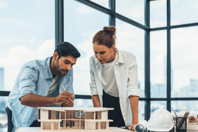 Duas pessoas analisando uma maquete arquitetônica sobre uma mesa, em um escritório com grandes janelas. Na mesa há capacete de segurança, rolos de papel e materiais de desenho.