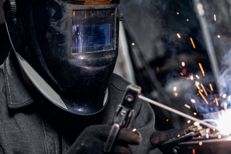 Pessoa utilizando máscara de solda enquanto realiza soldagem, com faíscas e luz intensa saindo do ponto de trabalho.