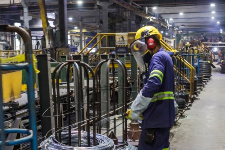 Ambiente industrial com máquinas e estruturas metálicas, onde um trabalhador usando capacete, protetor facial, luvas e uniforme de segurança opera entre bobinas de aço e equipamentos de produção.