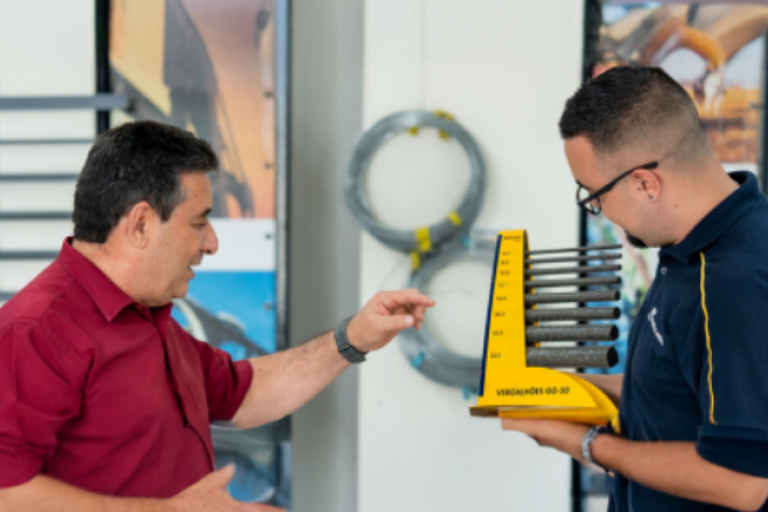 Duas pessoas interagem enquanto uma delas segura um mostruário amarelo com vergalhões de aço Gerdau em um ambiente de demonstração de produtos.