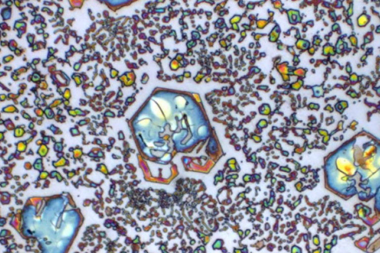 Imagem microscópica de metal com formas geométricas coloridas e partículas espalhadas, formando padrões visuais abstratos.