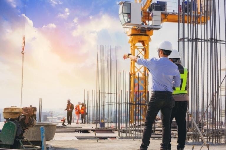 Dois profissionais usando capacetes de segurança estão em um canteiro de obras, observando e apontando para a estrutura de uma construção. No local há vergalhões verticais expostos e uma grande grua amarela ao fundo. A cena ocorre ao ar livre, com iluminação natural suave, sugerindo início ou fim de dia.