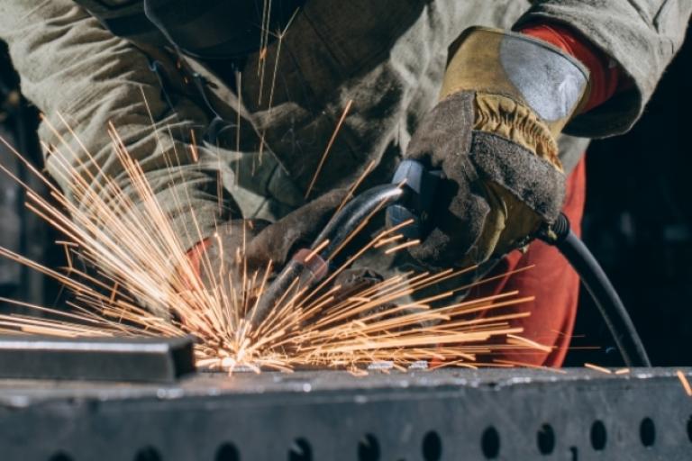Profissional de serralheria utilizando ferramenta elétrica para soldar ou desbastar uma peça de metal, com muitas faíscas sobre uma superfície de aço em ambiente industrial.