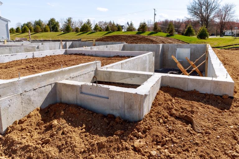Estrutura de fundação de uma casa em construção, composta por paredes de concreto formando diversos compartimentos subterrâneos, com terra ao redor e dentro das áreas delimitadas. No ambiente externo, há um terreno aberto com grama verde, árvores ao fundo, postes e fios elétricos, além de um céu claro com poucas nuvens. Dentro da fundação, aparecem suportes de madeira inclinados e pequenas aberturas na parede de concreto.