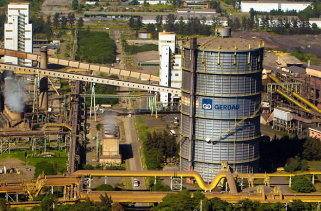 Vista aérea de um complexo industrial de siderurgia com estruturas metálicas, silos cilíndricos, tubulações e áreas de produção, com o logotipo da Gerdau em um dos grandes reservatórios.