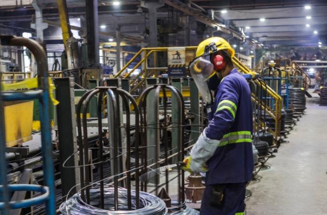 Ambiente industrial com máquinas e estruturas metálicas, onde um trabalhador usando capacete, protetor facial, luvas e uniforme de segurança opera entre bobinas de aço e equipamentos de produção.