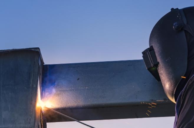 Pessoa utilizando equipamento de proteção para soldagem enquanto realiza solda utilizando eletrodo, produzindo faíscas e brilho intenso no ponto de contato