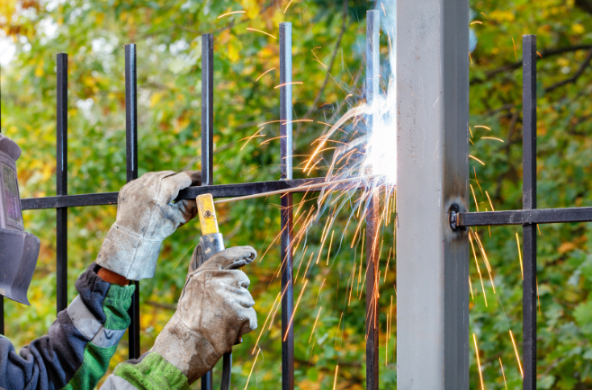 Pessoa usando luvas de proteção solda as barras de um portão metálico à sua estrutura, com faíscas visíveis, em ambiente externo com árvores ao fundo.