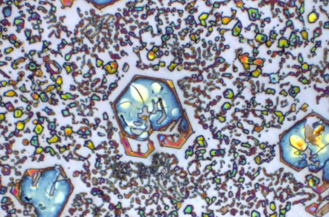 Imagem microscópica de metal com formas geométricas coloridas e partículas espalhadas, formando padrões visuais abstratos.