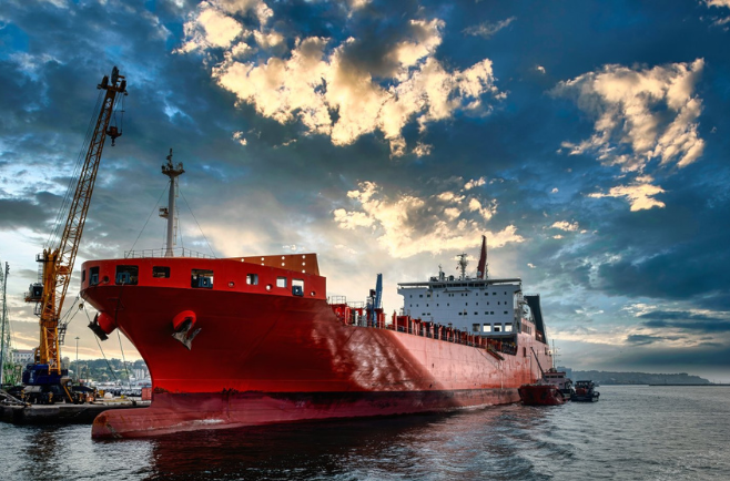Navio de carga vermelho em estaleiro com guindastes e estrutura industrial ao fundo