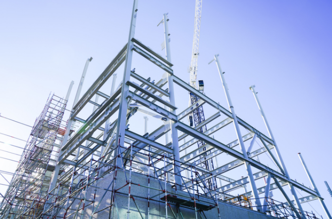 Estrutura metálica em construção com pilares e vigas de aço, andaimes e grua ao fundo.