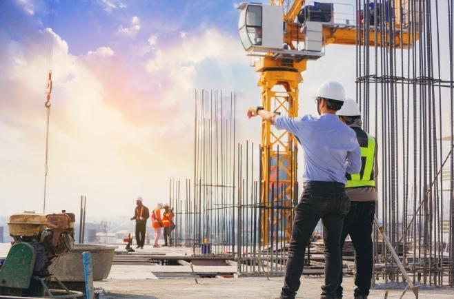 Dois profissionais usando capacetes de segurança estão em um canteiro de obras, observando e apontando para a estrutura de uma construção. No local há vergalhões verticais expostos e uma grande grua amarela ao fundo. A cena ocorre ao ar livre, com iluminação natural suave, sugerindo início ou fim de dia.