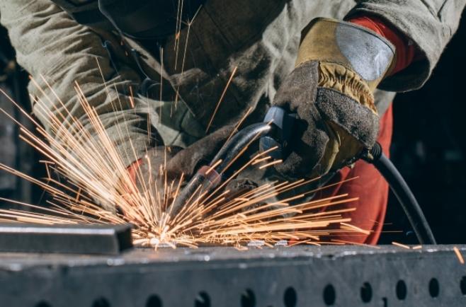 Profissional de serralheria utilizando ferramenta elétrica para soldar ou desbastar uma peça de metal, com muitas faíscas sobre uma superfície de aço em ambiente industrial.