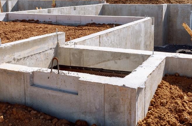 Estrutura de fundação de uma casa em construção, composta por paredes de concreto formando diversos compartimentos subterrâneos, com terra ao redor e dentro das áreas delimitadas. No ambiente externo, há um terreno aberto com grama verde, árvores ao fundo, postes e fios elétricos, além de um céu claro com poucas nuvens. Dentro da fundação, aparecem suportes de madeira inclinados e pequenas aberturas na parede de concreto.