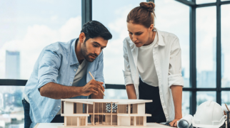 Duas pessoas analisando uma maquete arquitetônica sobre uma mesa, em um escritório com grandes janelas. Na mesa há capacete de segurança, rolos de papel e materiais de desenho.