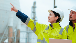 Dois profissionais com capacetes e coletes de segurança analisando uma área industrial com grandes tanques esféricos ao fundo, um deles segurando um laptop e apontando para a estrutura.