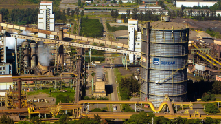 Vista aérea de um complexo industrial de siderurgia com estruturas metálicas, silos cilíndricos, tubulações e áreas de produção, com o logotipo da Gerdau em um dos grandes reservatórios.