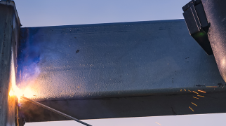 Pessoa utilizando equipamento de proteção para soldagem enquanto realiza solda utilizando eletrodo, produzindo faíscas e brilho intenso no ponto de contato