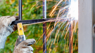 Pessoa usando luvas de proteção solda as barras de um portão metálico à sua estrutura, com faíscas visíveis, em ambiente externo com árvores ao fundo.