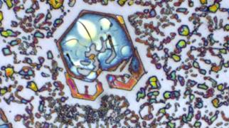 Imagem microscópica de metal com formas geométricas coloridas e partículas espalhadas, formando padrões visuais abstratos.