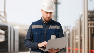 Profissional em ambiente industrial usando capacete e colete azul escuro, operando um notebook entre estruturas metálicas.