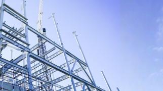 Estrutura metálica em construção com pilares e vigas de aço, andaimes e grua ao fundo.