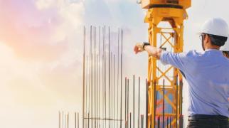 Dois profissionais usando capacetes de segurança estão em um canteiro de obras, observando e apontando para a estrutura de uma construção. No local há vergalhões verticais expostos e uma grande grua amarela ao fundo. A cena ocorre ao ar livre, com iluminação natural suave, sugerindo início ou fim de dia.