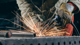 Profissional de serralheria utilizando ferramenta elétrica para soldar uma peça de metal, com muitas faíscas sobre uma superfície de aço em ambiente industrial.