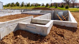 Estrutura de fundação de uma casa em construção, composta por paredes de concreto formando diversos compartimentos subterrâneos, com terra ao redor e dentro das áreas delimitadas. No ambiente externo, há um terreno aberto com grama verde, árvores ao fundo, postes e fios elétricos, além de um céu claro com poucas nuvens. Dentro da fundação, aparecem suportes de madeira inclinados e pequenas aberturas na parede de concreto.